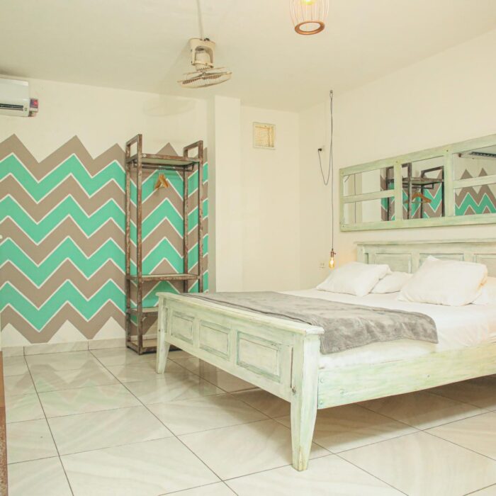 Habitación con cama grande, paredes decoradas con patrón de chevron en tonos verdes y grises, mobiliario de madera en estilo shabby chic, aire acondicionado y lámparas de ambiente.