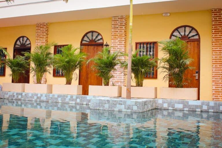 Masaya-experience-hotel-piscina-borde-piscina-cafe-masaya- Experience hotel with vibrant yellow walls, wooden doors, and lush green plants in decorative planters surrounding a refreshing swimming pool. Perfect for relaxing stays.