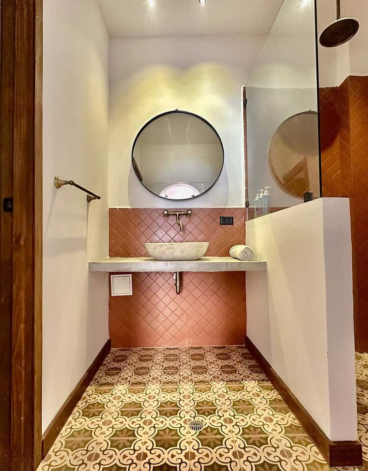 Esquina de baño con lavamanos de piedra, espejo redondo y azulejos terracota en pared y suelo con diseño clásico, estilo moderno y decoración elegante para una experiencia de alojamiento en Masaya.