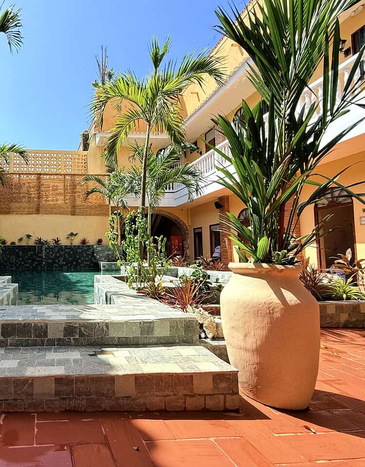 Un patio con piscina en un entorno tropical, rodeado de palmas y macetas de barro. El área combina elementos de piedra y cerámica, creando un espacio relajante y elegante para relajarse y disfrutar del sol.