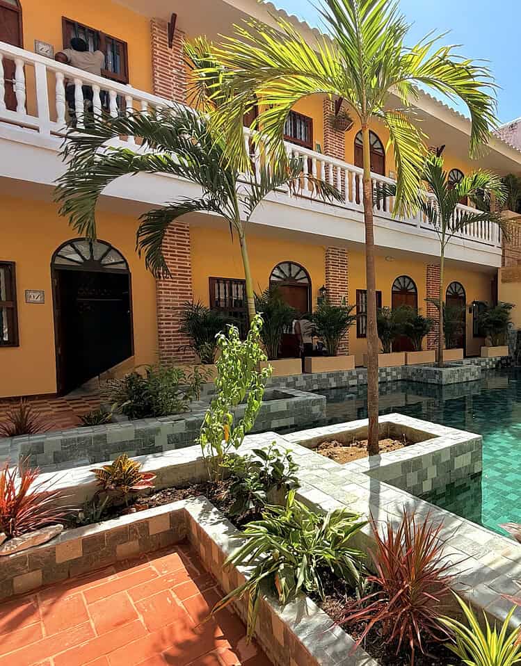 Pisos de color terracota y paredes amarillas rodeadas de palmeras y plantas verdes en un hotel boutique con piscina central, bajo un cielo azul claro ideal para vacaciones y relajación.
