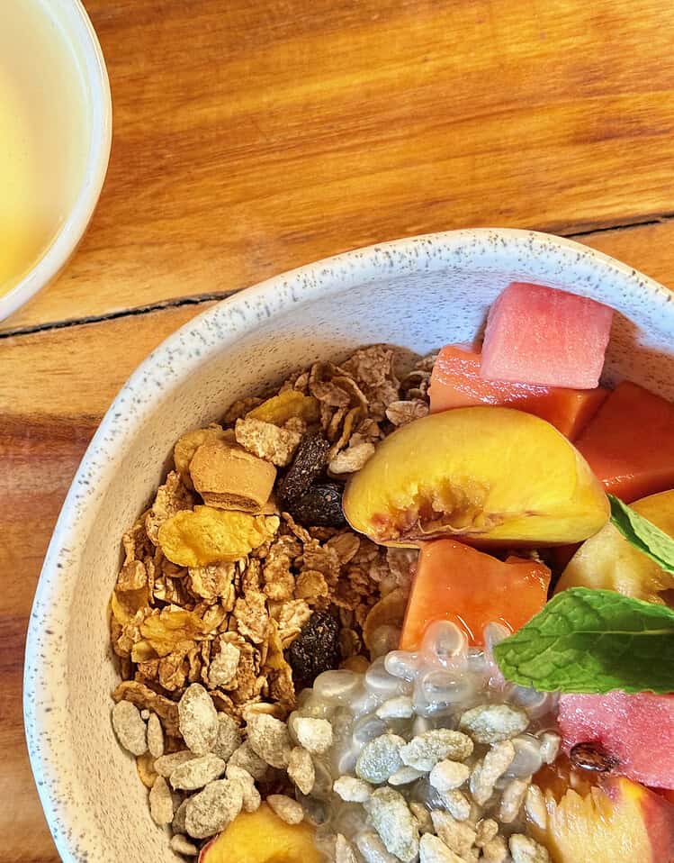 Arroz con frutas, granola y semillas en un bowl, acompañado de jugo de mango en una taza blanca sobre una mesa de madera. Una opción saludable y nutritiva para comenzar el día.