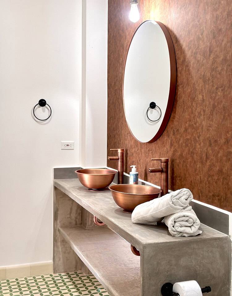 Baño moderno con lavabos de cobre, espejo ovalado y toallas enrolladas en un mueble de concreto.