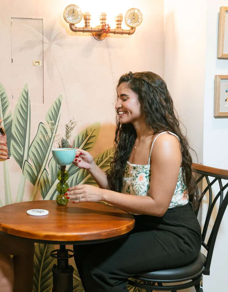 Dos mujeres sentadas en una mesa de madera, compartiendo una bebida en un ambiente acogedor y decorado con plantas y arte en las paredes.