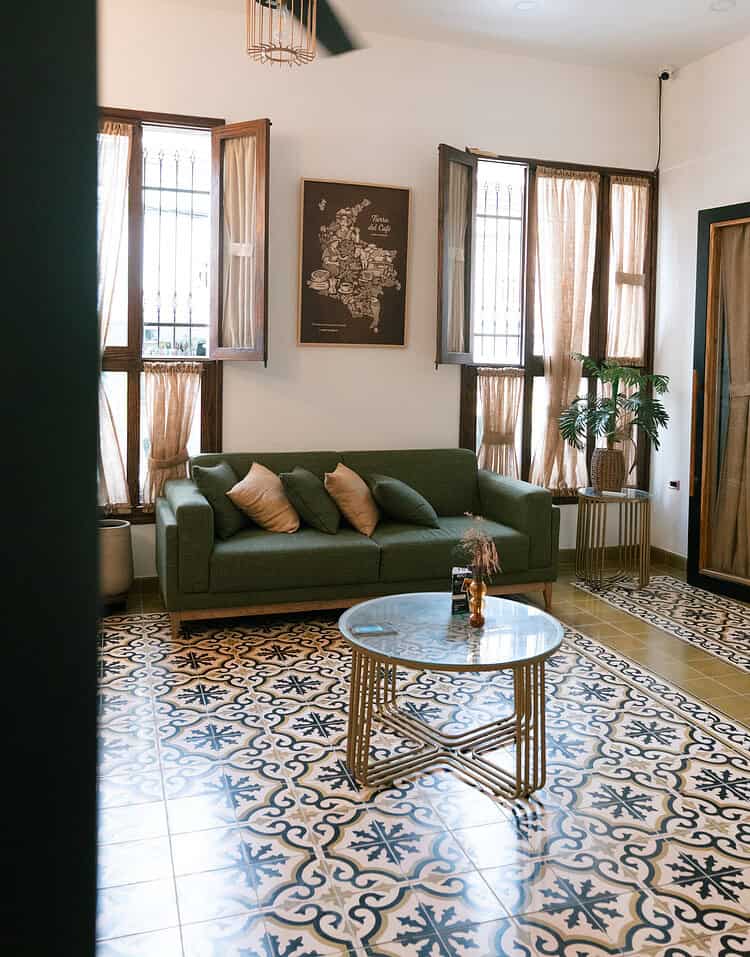 Un espacio acogedor con sofá verde, cojines y una mesa de centro en una sala con ventanas grandes y cortinas de tela ligera.