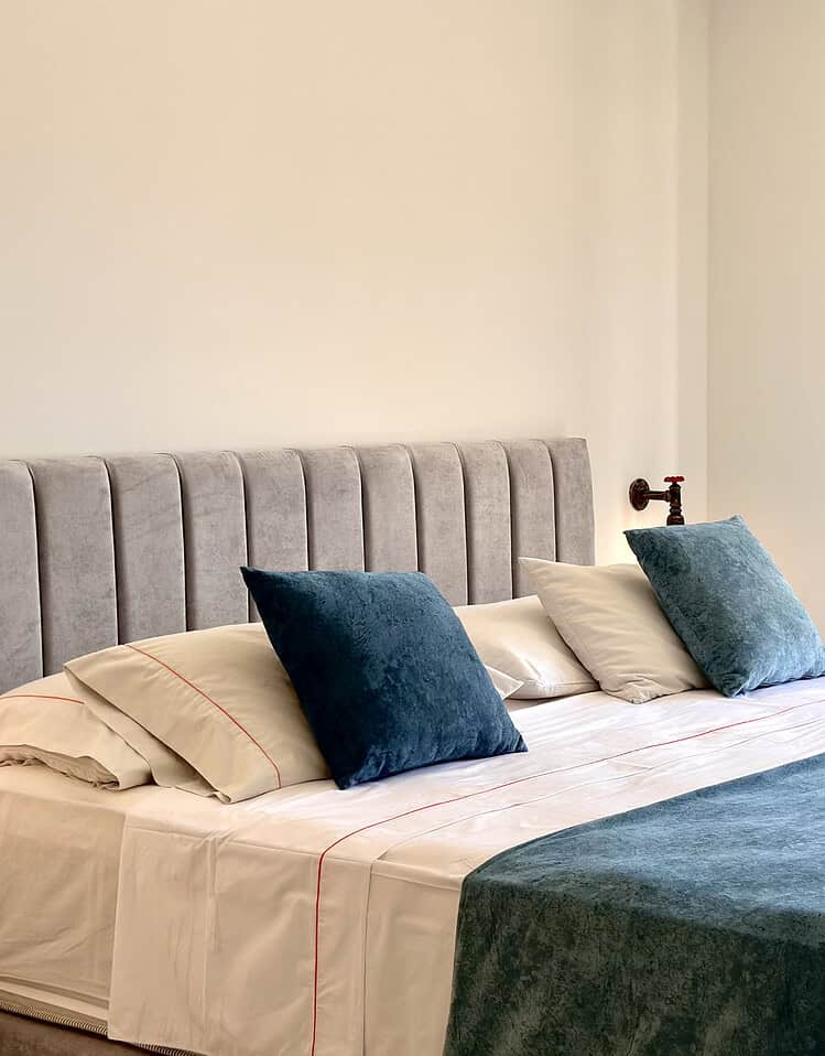 Una habitación de hotel con cama doble, almohadas y ropa de cama en tonos neutros y azules, con una ventana grande que permite la entrada de luz natural y ofrece vista a la ciudad.