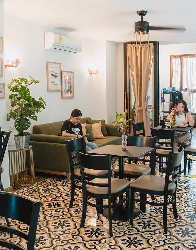 Interior del comedor en un hotel boutique con ambiente cálido y relajado, con varias mesas y sillas, plantas decorativas y paredes con cuadros, ideal para disfrutar de una comida o café.