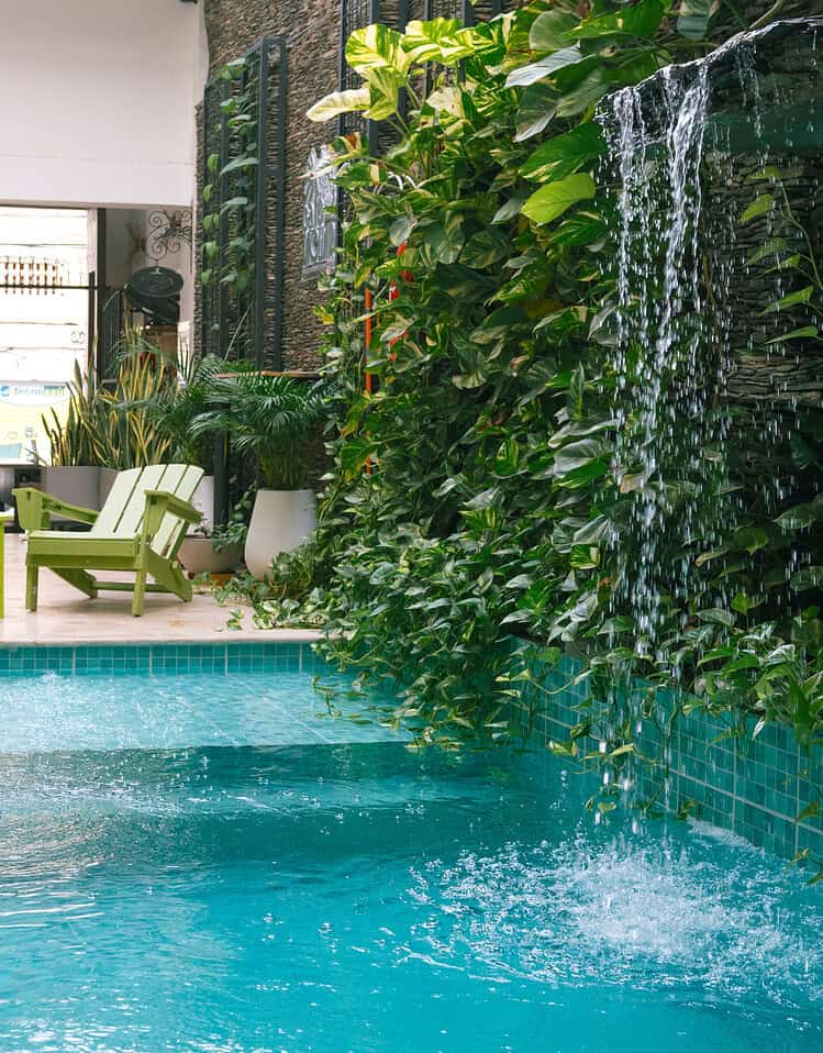 Piscina con cascada y zona de descanso con sillas verdes y plantas tropicales. Ambiente relajante y natural en un hotel boutique de Masaya.