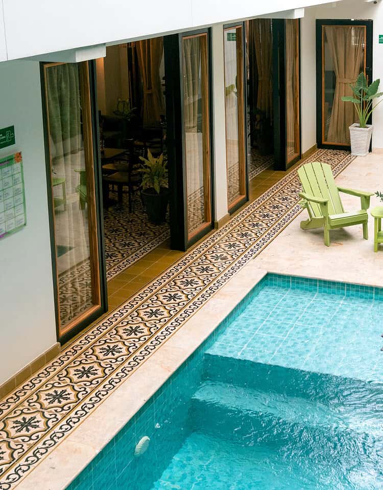 Piscina en un hotel boutique con área de descanso con sillas verdes y plantas, rodeada de habitaciones con puertas de vidrio, creando un ambiente relajante y acogedor.