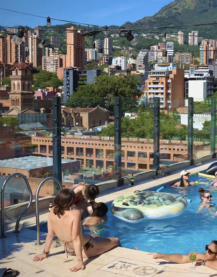 Personas disfrutando en la piscina en la terraza con vista a la ciudad y montañas en el fondo.