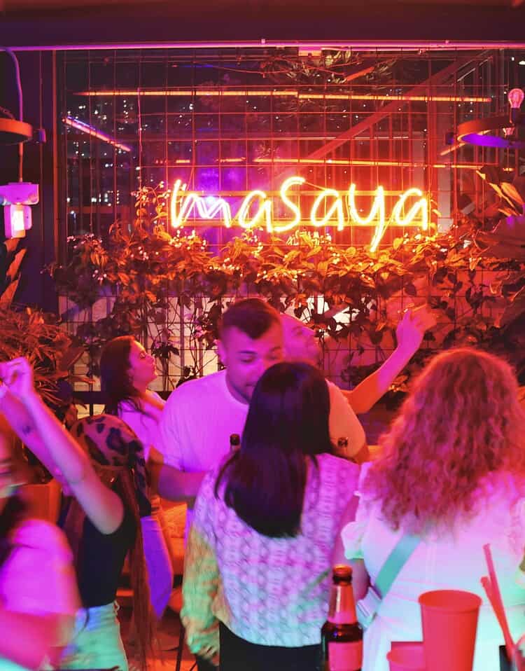 Grupo de personas disfrutando en un bar con iluminación rosa y neón que dice 'masaya' en el fondo.