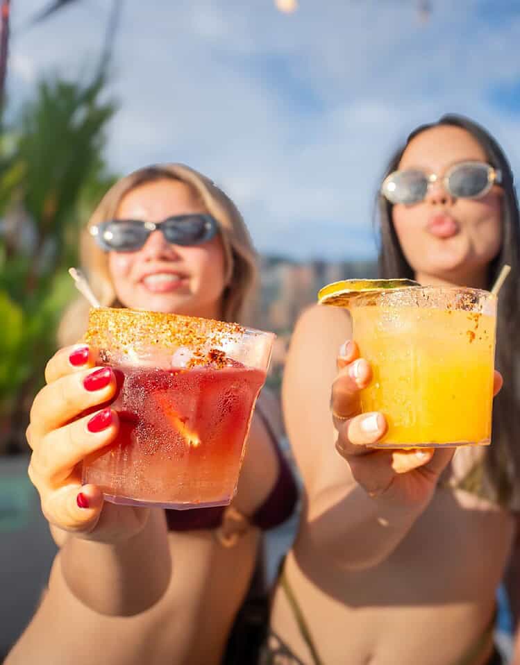 Tres mujeres con gafas de sol disfrutando cócteles en la playa bajo un cielo azul.