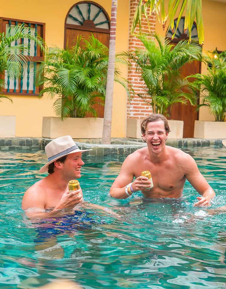 Tres amigos disfrutando en la piscina, sonriendo y tomando bebidas en un ambiente tropical y relajado en M.