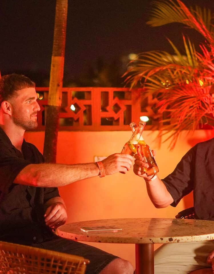 Dos jóvenes brindando con bebidas en una terraza con iluminación cálida y plantas tropicales.