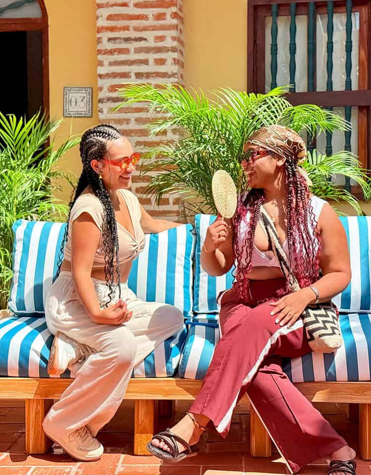 Dos mujeres conversando y riendo en un patio con plantas verdes y muebles de madera en un hotel de M.