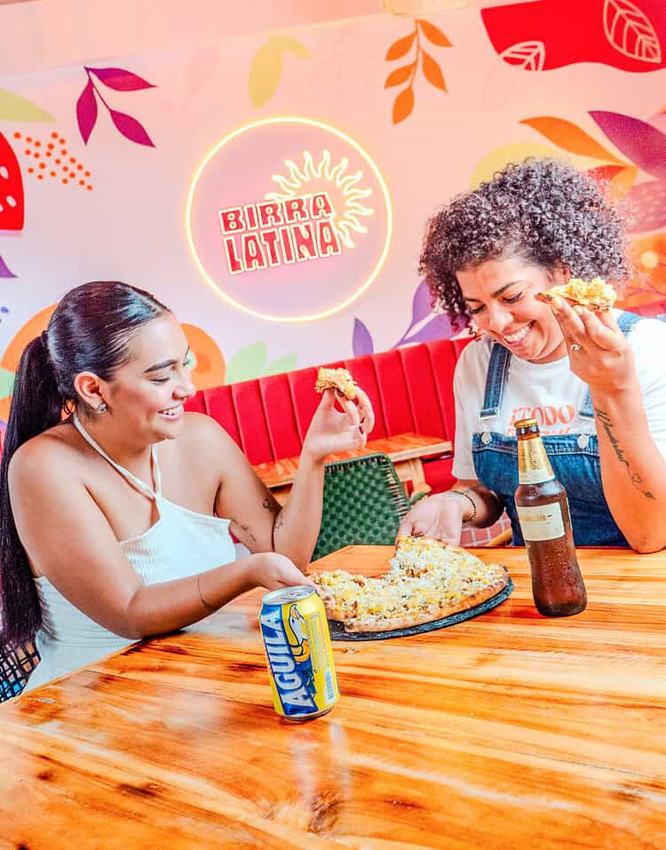 Dos mujeres disfrutando pizza en un restaurante con decoración colorida y ambiente alegre.