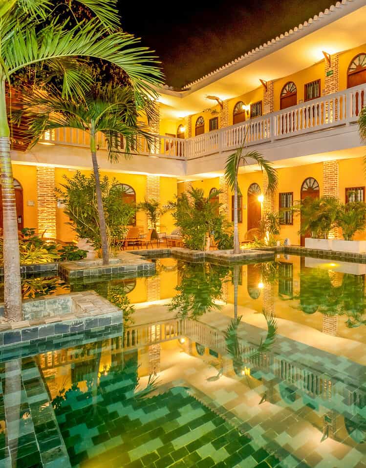Vista nocturna de la piscina y el hotel con iluminación cálida y plantas tropicales.