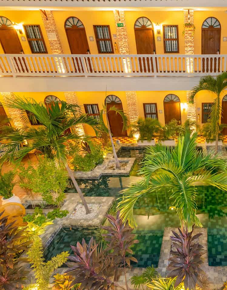 Vista del patio de M con piscina, jardines tropicales y arquitectura colonial iluminada de noche.