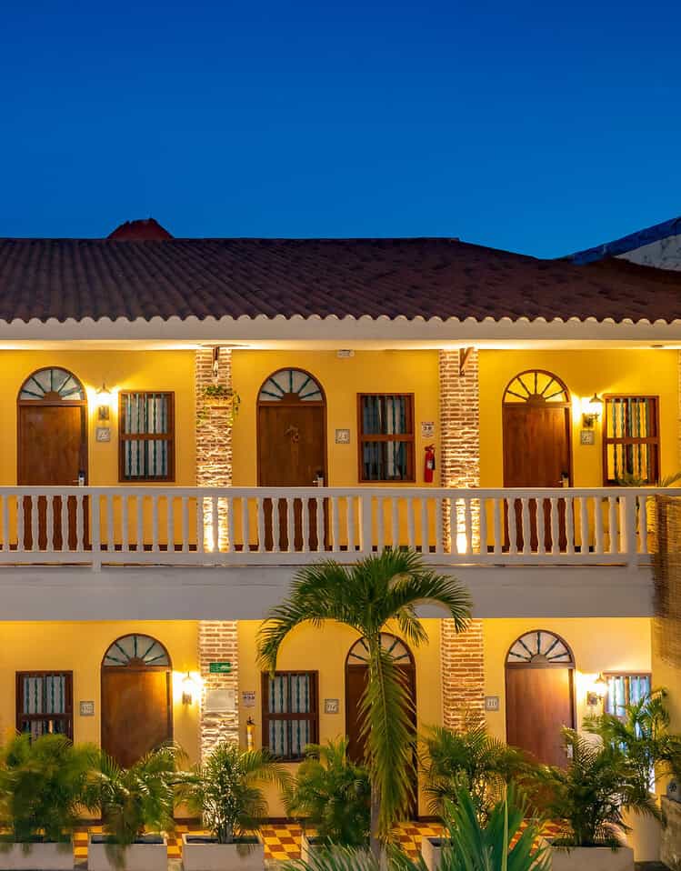 Fachada de hotel colonial iluminada en la noche con balcones y plantas tropicales en primer plano.