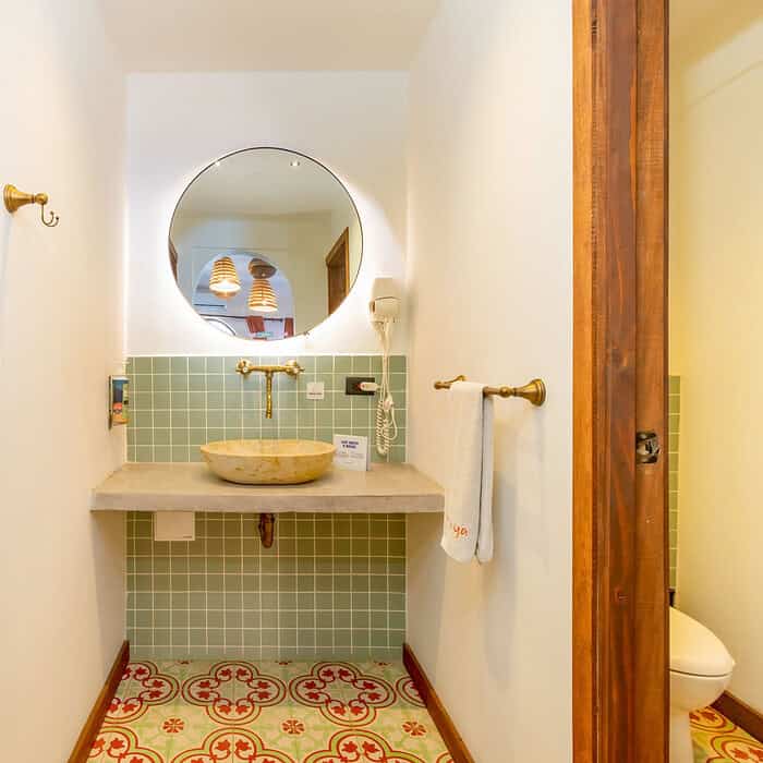 Baño con paredes de azulejos verdes y piso decorativo, lavabo de piedra y espejo redondo.