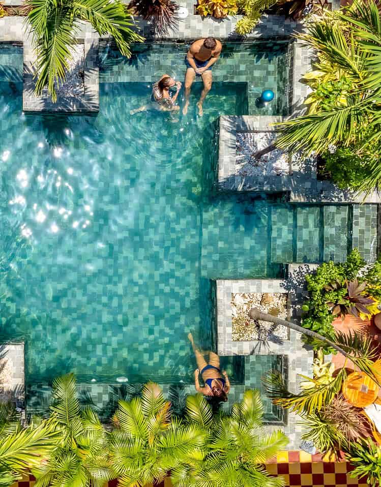 Piscina en M con áreas de descanso y personas disfrutando del agua en un entorno tropical.