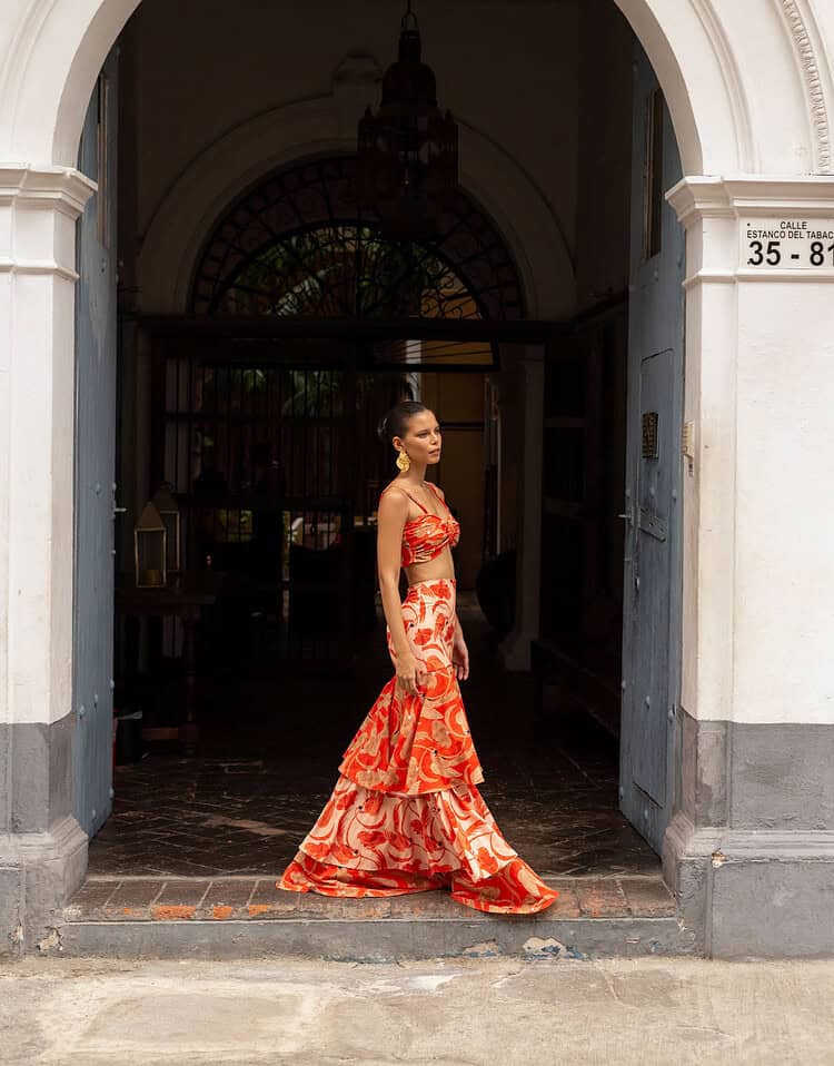 Mujer con vestido largo y estampado en tonos rojos y naranjas en la entrada de un edificio colonial.