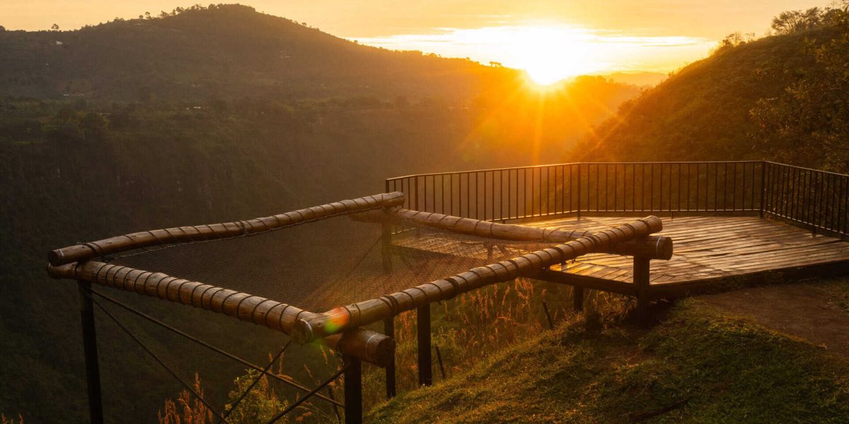Mirador de madera con barandal, sobre un barranco montañoso, durante un atardecer dorado que ilumina la escena y resalta la belleza natural del paisaje.